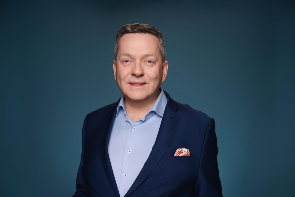 Andre Hoppen - Fotorealistisches Portrait eines lächelnden Mannes im schwarzen Anzug und weißem Hemd vor blauem Hintergrund (KI-generiert)