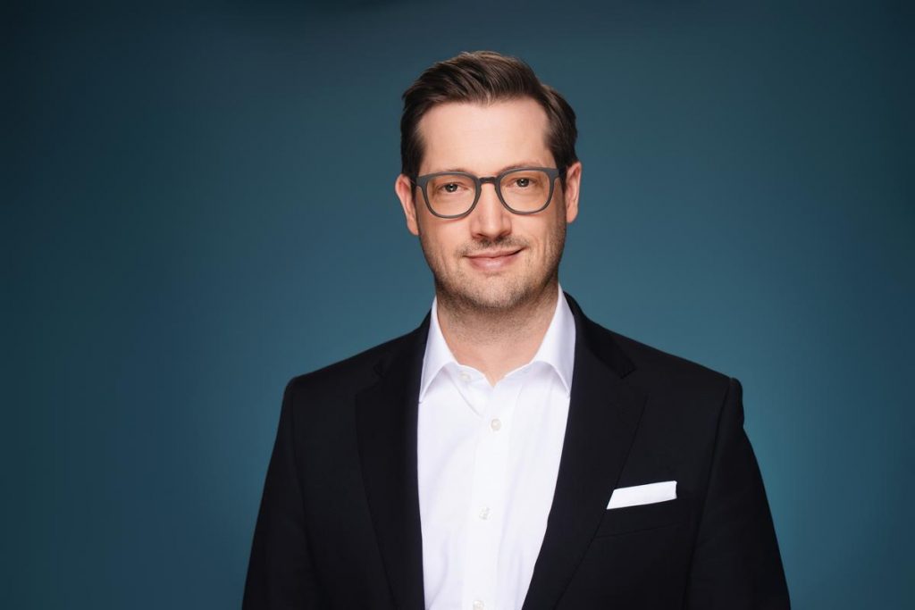 Peter Goldbach - Fotorealistisches Portrait eines lächelnden Mannes im schwarzen Anzug und weißem Hemd vor blauem Hintergrund (KI-generiert)