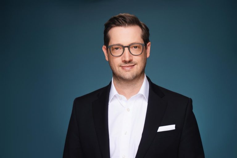 Peter Goldbach - Fotorealistisches Portrait eines lächelnden Mannes im schwarzen Anzug und weißem Hemd vor blauem Hintergrund (KI-generiert)